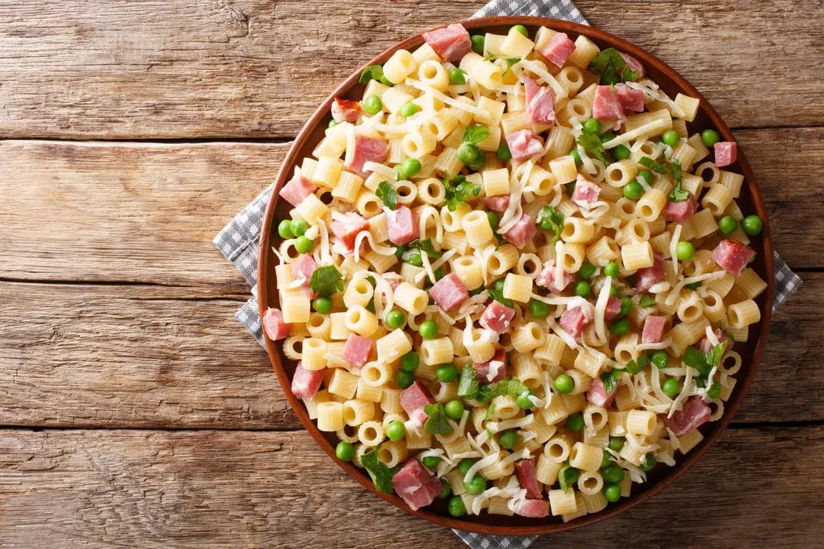 Un piatto di pasta con prosciutto cotto, piselli e formaggio grattugiato su un tavolo di legno rustico.