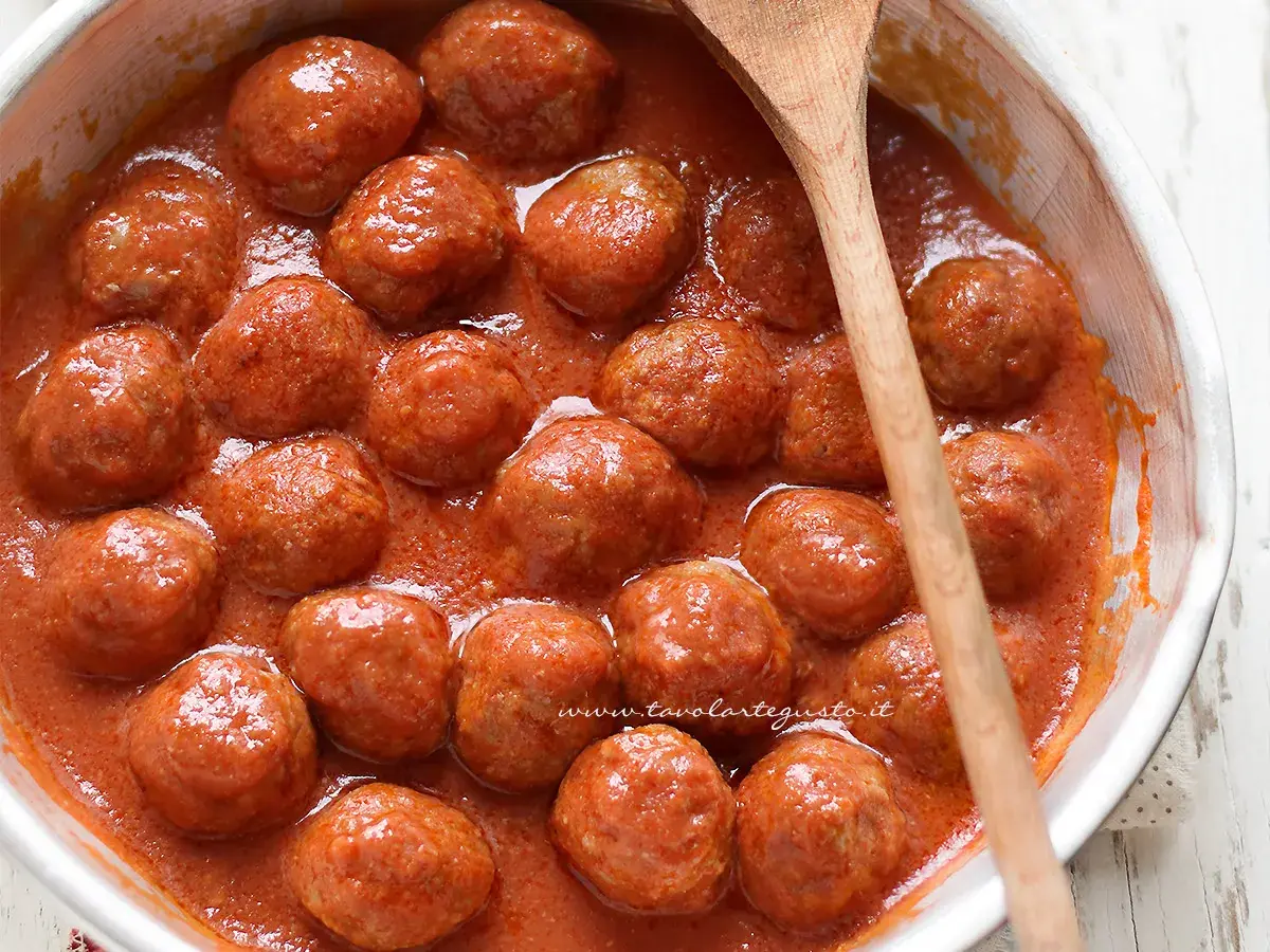 Polpette al sugo succulente in una ciotola bianca, pronte per essere gustate. Un cucchiaio di legno le accompagna.