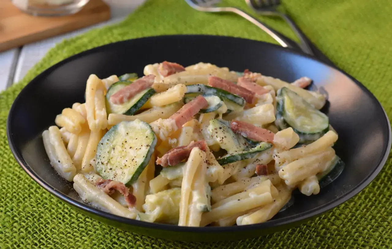 Piatto di pasta cremosa con zucchine a rondelle e cubetti di speck croccante, guarnito con pepe nero macinato.