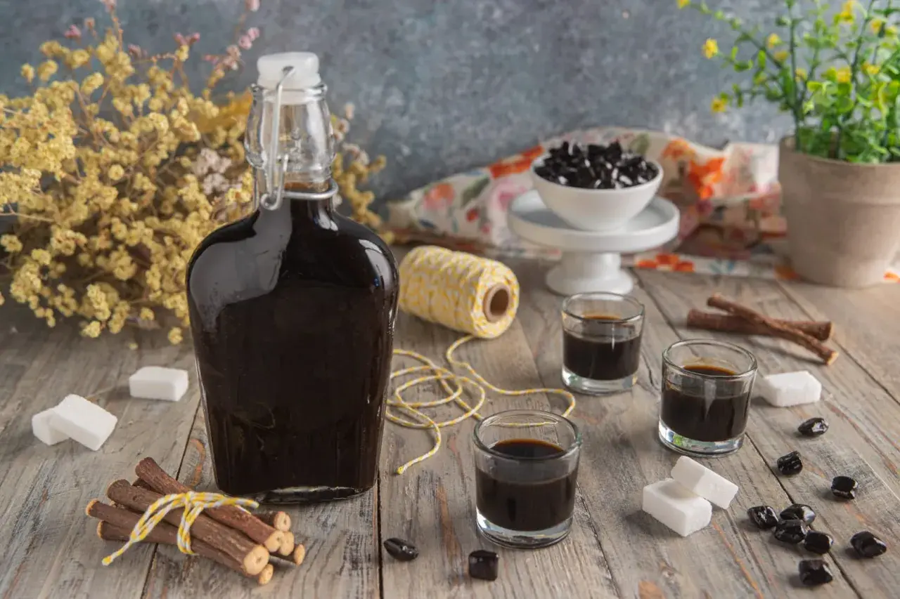 Bottiglia di liquore alla liquirizia scuro, bicchierini, bastoncini di liquirizia, zollette di zucchero e caramelle nere su un tavolo di legno.
