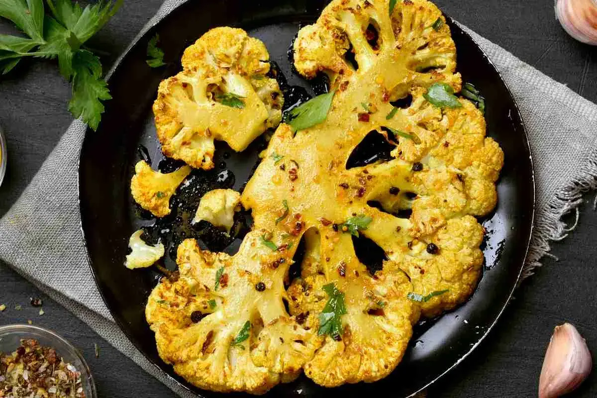 Fette di cavolo al forno dorate e speziate, servite su una padella nera con erbe fresche e pepe nero.