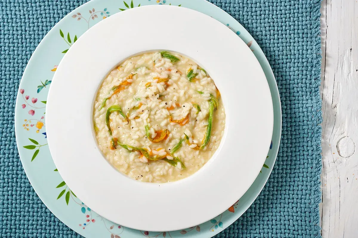 Un piatto di risotto ai fiori di zucca cremoso, con petali arancioni e fili verdi che si intrecciano nel riso bianco.
