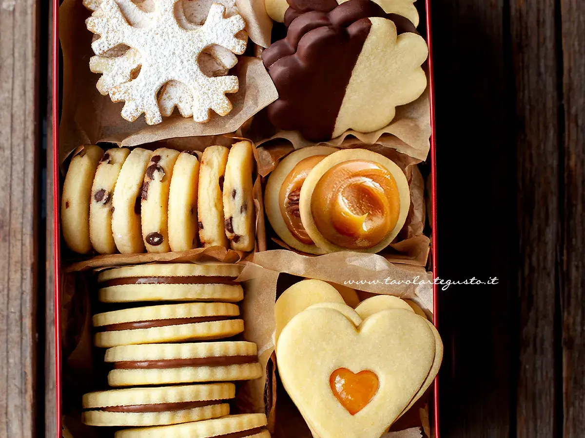 Scatola piena di biscotti classici: a fiocco di neve, con gocce di cioccolato, ripieni di caramello, a forma di cuore e ricoperti di cioccolato.
