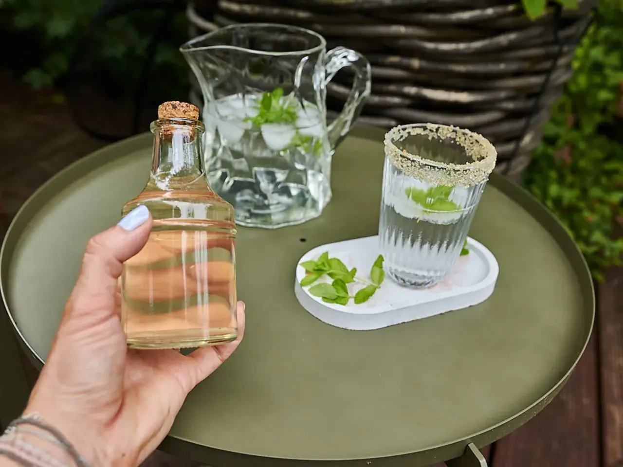 Mano tiene una bottiglietta di sciroppo di menta, accanto a un bicchiere e una caraffa con acqua, ghiaccio e foglie di menta.