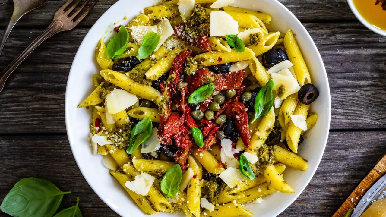 Penne al pesto con pomodori secchi, olive nere, capperi e scaglie di parmigiano. Un piatto colorato e saporito.