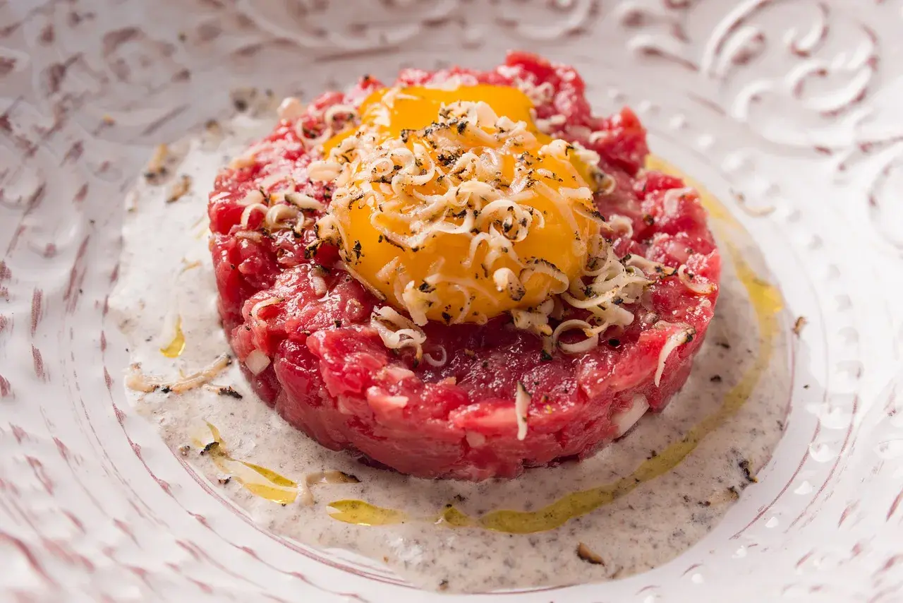 Deliziosa tartare di manzo finemente tritata, guarnita con tuorli d'uovo dorati e scaglie di formaggio, servita su un piatto elegante con una salsa cremosa.