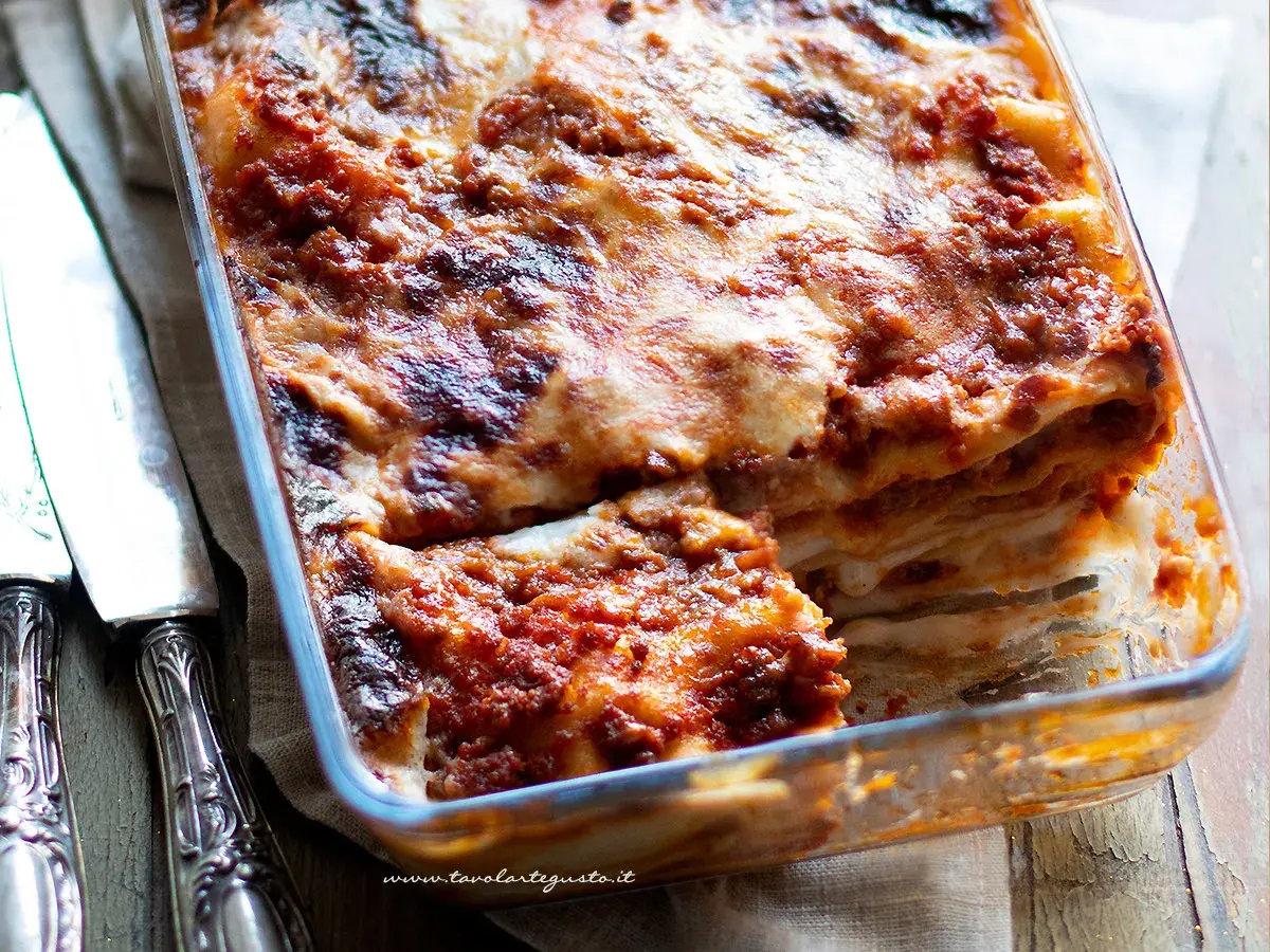 Lasagne alla bolognese appena sfornate, dorate e filanti, in una teglia di vetro. Accanto, due coltelli d'argento.