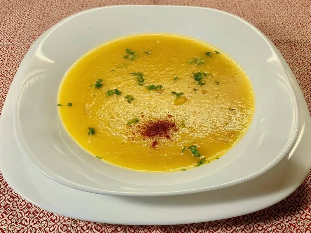 Una vellutata crema di peperoni, guarnita con prezzemolo fresco e una spolverata di paprika, servita in una ciotola bianca su un tovagliolo rosso.