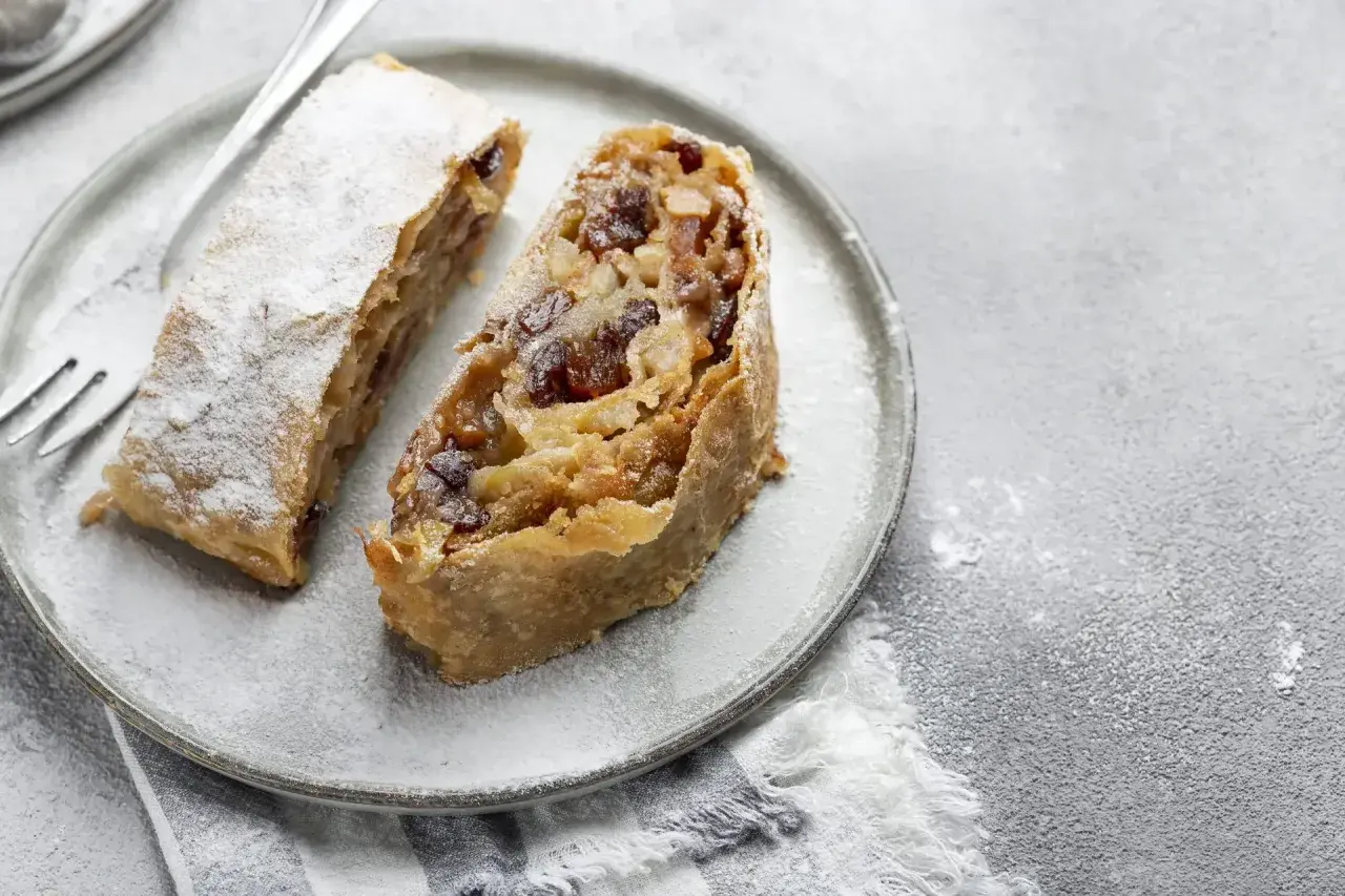 Due fette di strudel di mele con pasta sfoglia, cosparse di zucchero a velo, su un piatto grigio con una forchetta.
