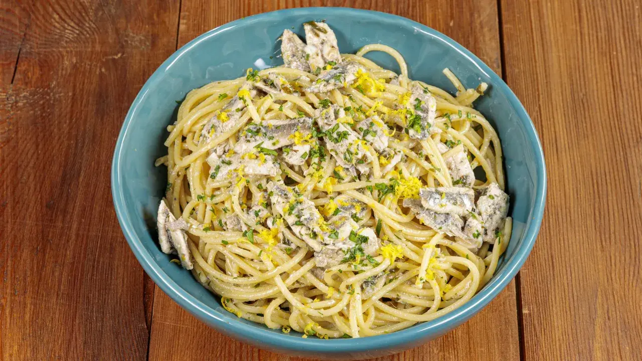 Spaghetti con acciughe, prezzemolo tritato e scorza di limone grattugiata in una ciotola blu su un tavolo di legno.