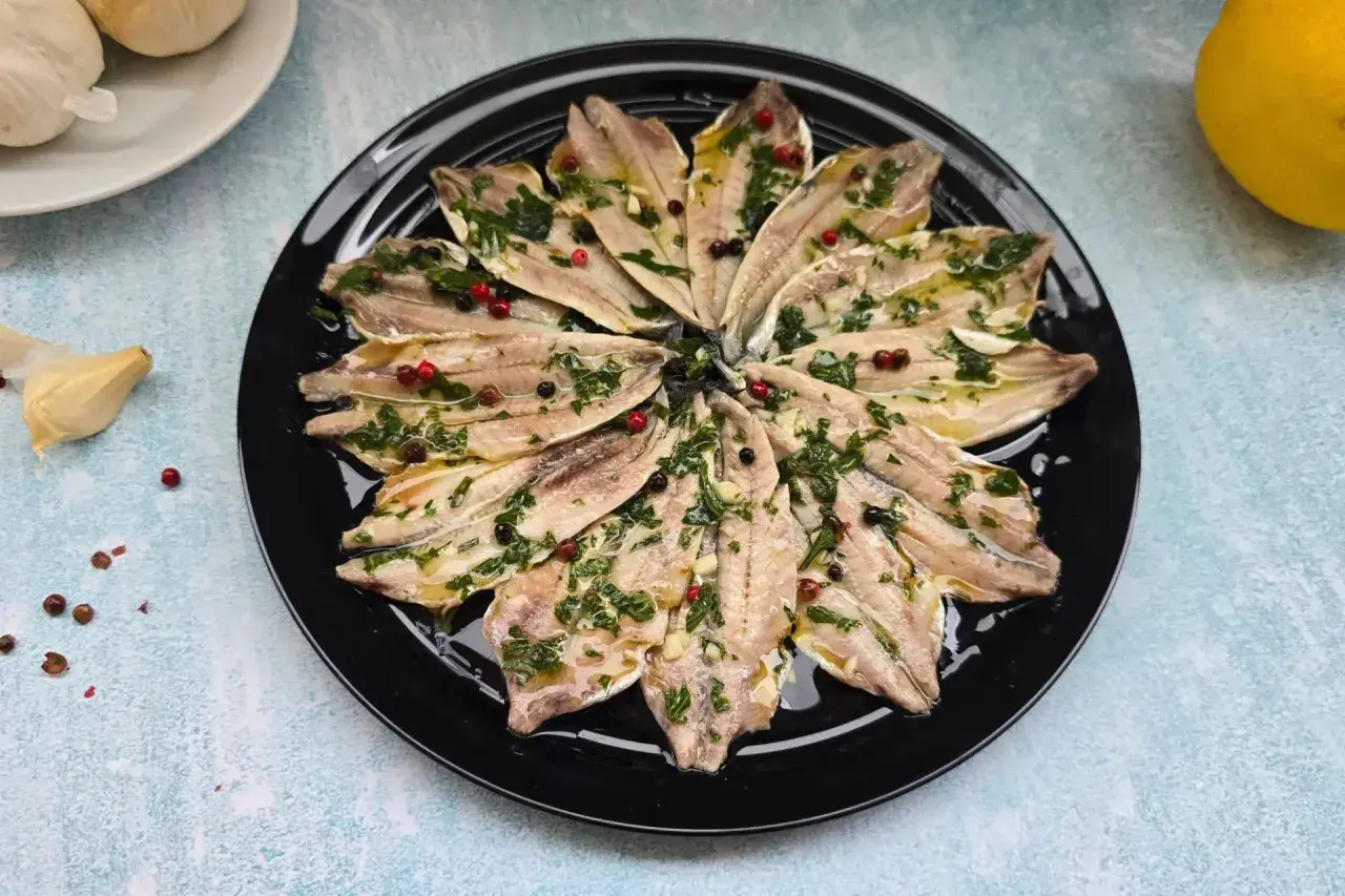 Alici marinate ricetta: filetti di acciughe conditi con erbe aromatiche, aglio e pepe in grani su un piatto nero.