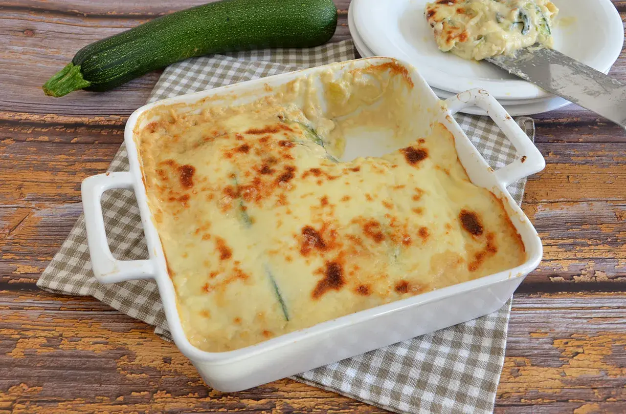 Parmigiana di zucchine bianca senza besciamella, dorata e filante, pronta per essere gustata. Accanto, una zucchina fresca.