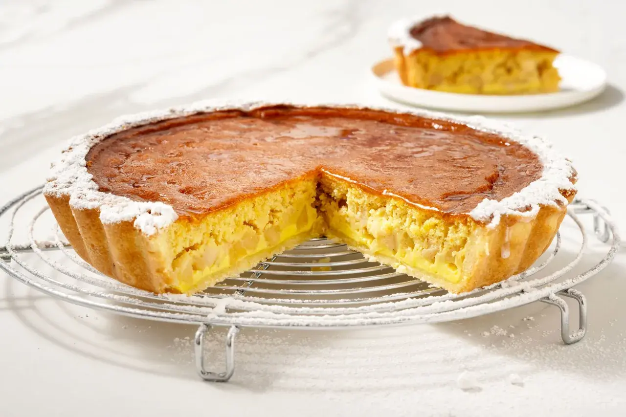 Torta di mele con crema pasticcera, con una fetta tagliata, cosparsa di zucchero a velo su una griglia metallica.