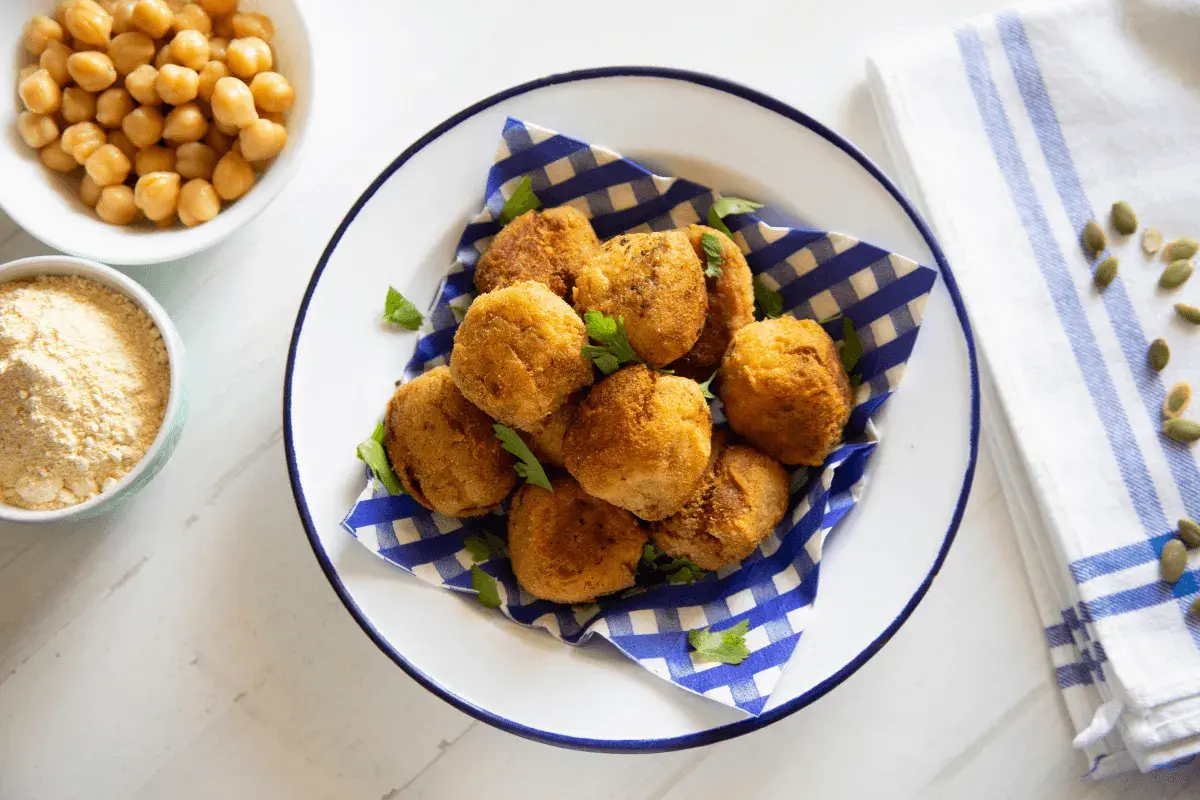 Piatto di polpette di ceci dorate e croccanti, guarnite con prezzemolo fresco. Accanto, ciotole con ceci e farina di ceci.