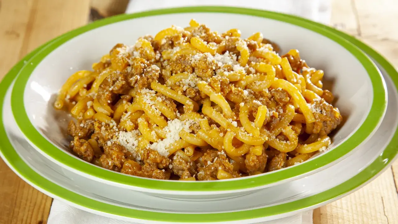 Piatto di gramigna alla salsiccia, condita con ragù e parmigiano grattugiato, servita in una ciotola bianca con bordo verde.