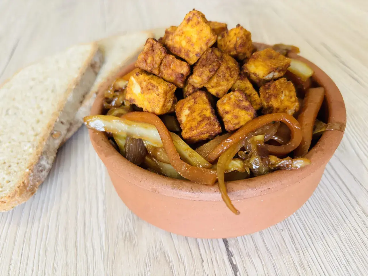 Tofu in padella dorato e saporito, servito con cipolle caramellate in una ciotola di terracotta, accanto a fette di pane rustico.