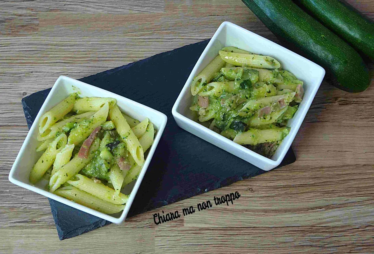 Due ciotole di pasta penne con zucchine e pancetta, condite con un pesto verde. Due zucchine fresche riposano sullo sfondo.