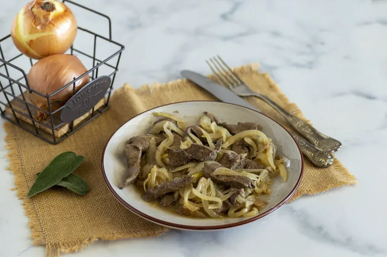 Piatto di fegato alla veneziana con cipolle dorate, accompagnato da cipolle fresche in un cestino e foglie di salvia.