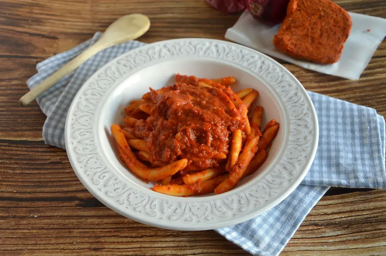 Piatto di pasta con sugo rosso piccante, condita con nduja calabrese. Accanto, un pezzo di nduja e cipolla rossa.
