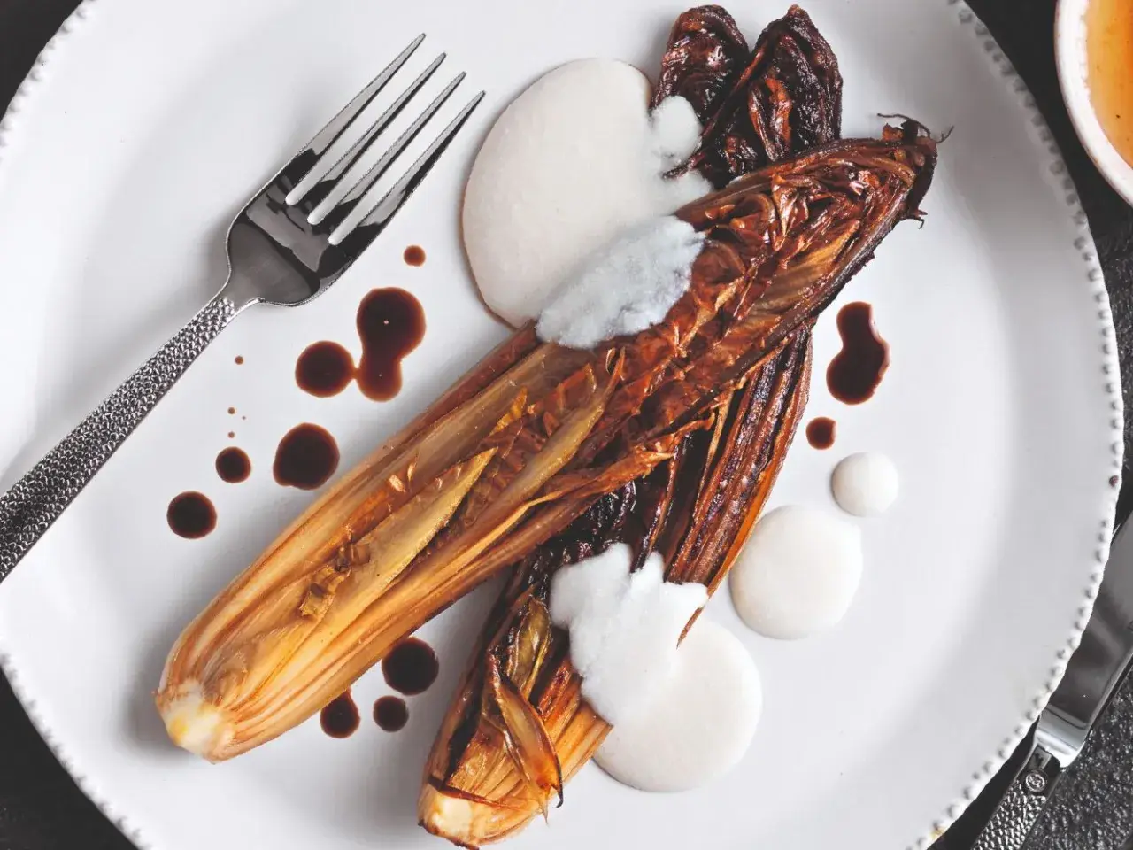 Radicchio al forno caramellato, servito con crema bianca e salsa scura su un piatto bianco, con forchetta e coltello.