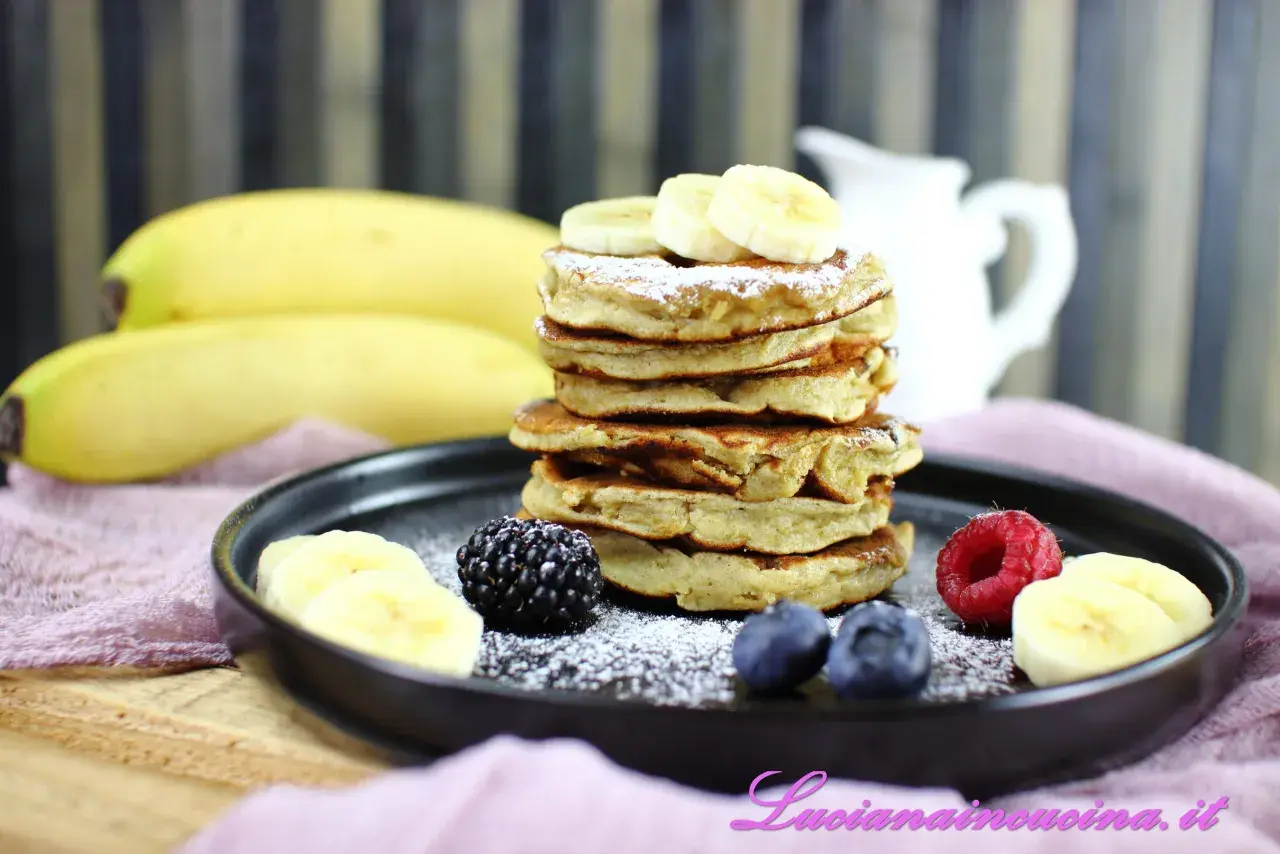 Pancakes alla banana soffici e dorati, guarniti con fettine di banana, frutti di bosco e zucchero a velo.