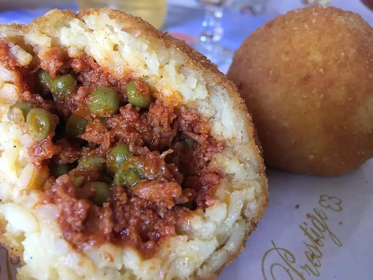Un arancino o arancina, tagliato a metà, rivela un ripieno di ragù e piselli. Accanto, un altro intero, dorato e croccante.