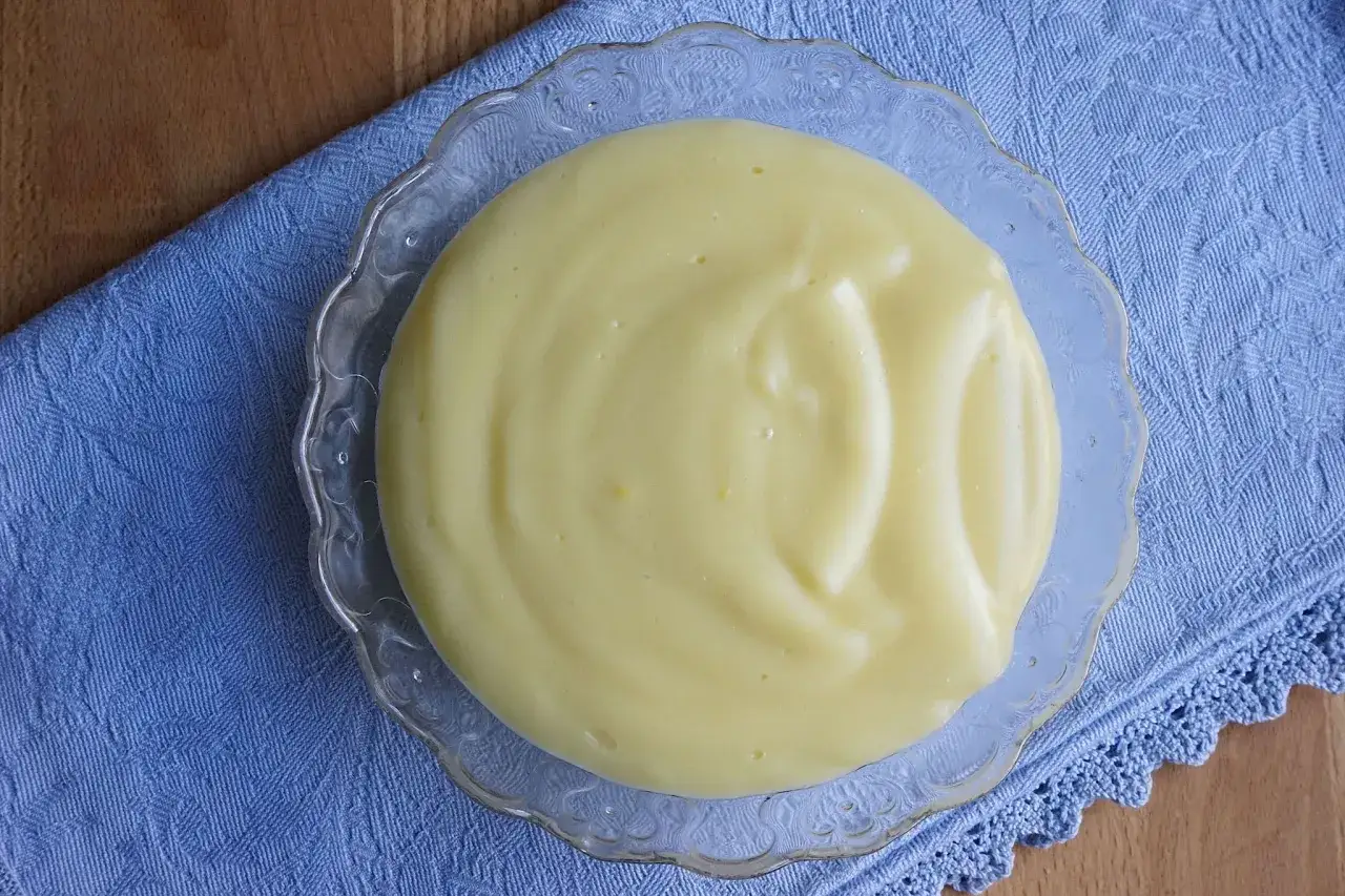 Crema pasticcera bimby, liscia e vellutata, servita in una ciotola di vetro su un tovagliolo blu.
