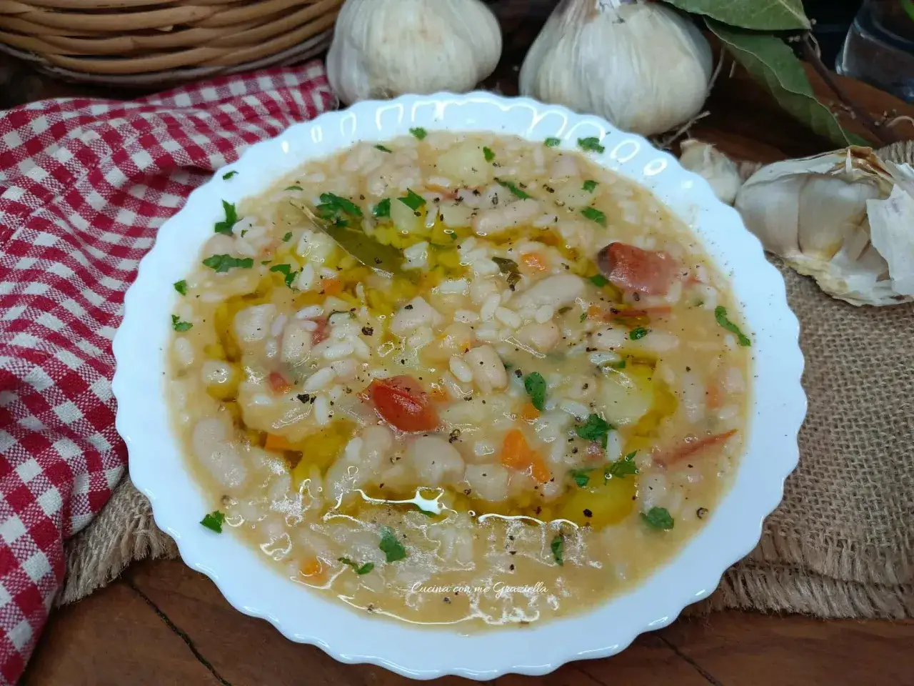 Piatto di riso e fagioli cremoso, condito con olio d'oliva, pomodorini e prezzemolo. Sullo sfondo, aglio e una tovaglia a quadri rossi.