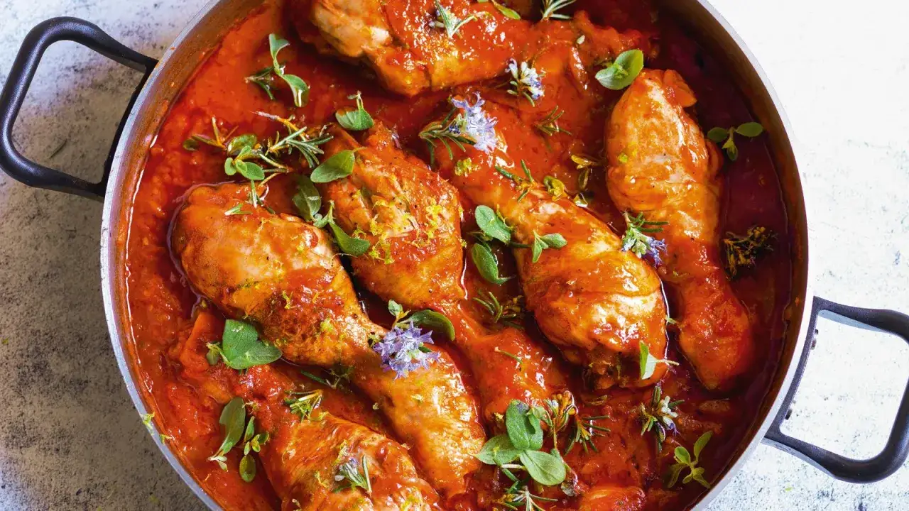 Un saporito pollo alla cacciatora in padella, con pezzi di pollo succulenti immersi in una ricca salsa rossa, guarnito con erbe aromatiche fresche e scorza di limone.