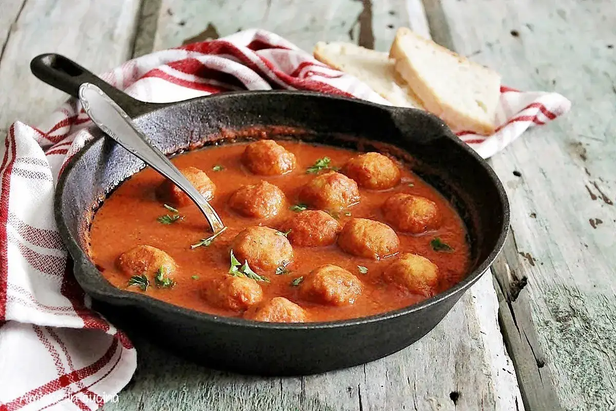 Polpette di pane in salsa di pomodoro, servite in una padella di ghisa con pane tostato e un tovagliolo a quadri rossi.