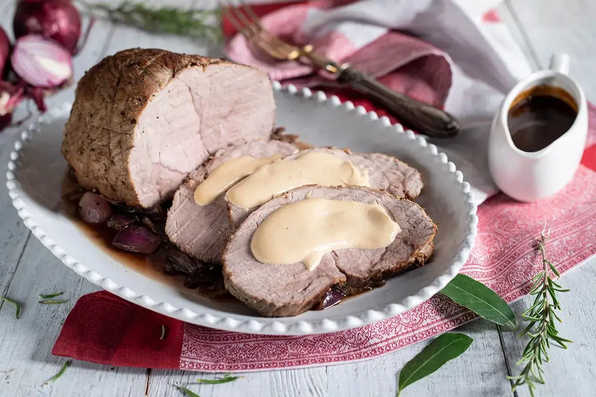 Arrosto di vitello morbido con sughetto benedetta, servito su un piatto bianco con cipolle rosse e rosmarino.