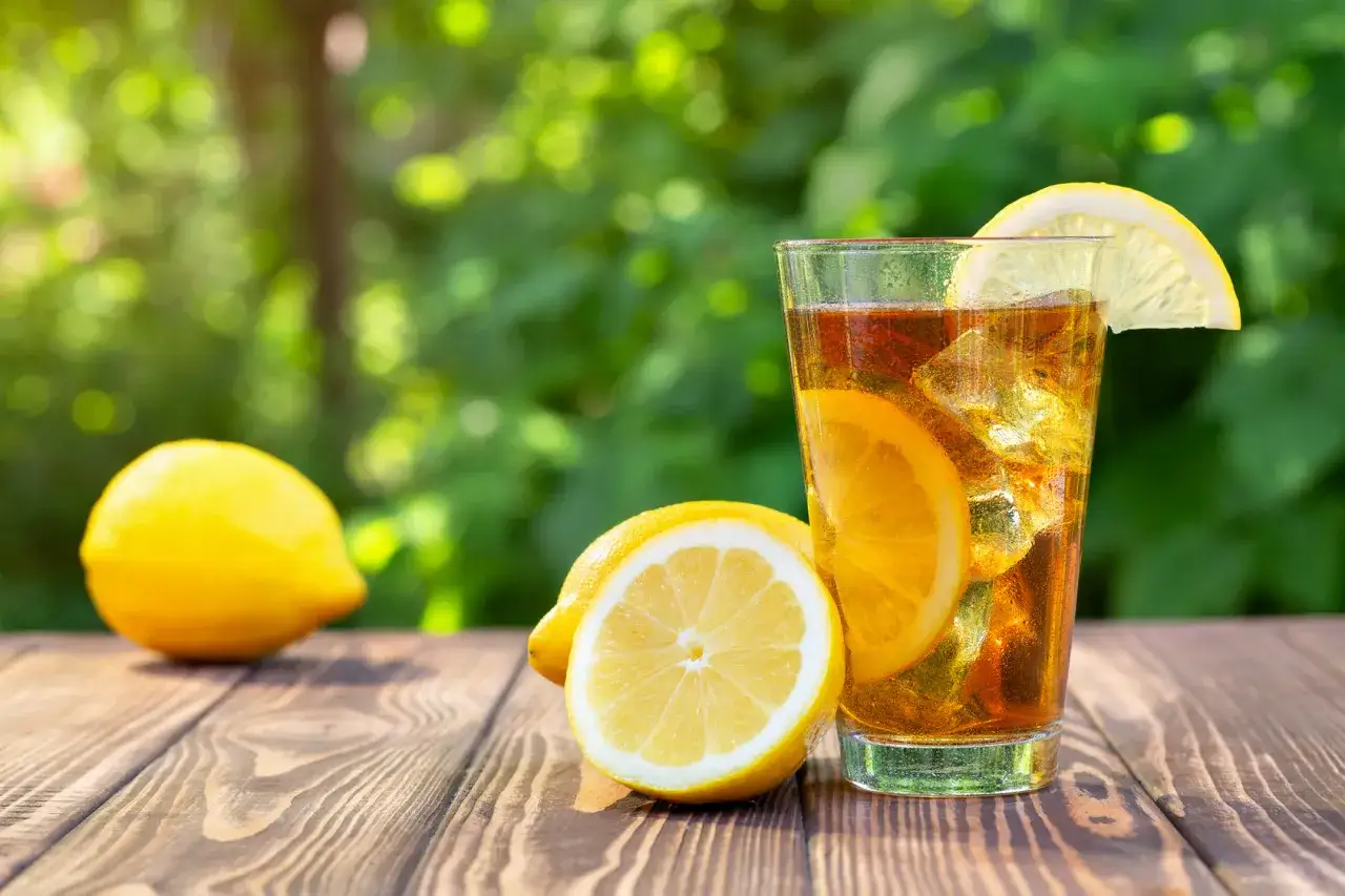 Un bicchiere di t&egrave; freddo fatto in casa con limone e ghiaccio, accompagnato da limoni freschi su un tavolo di legno all'aperto.