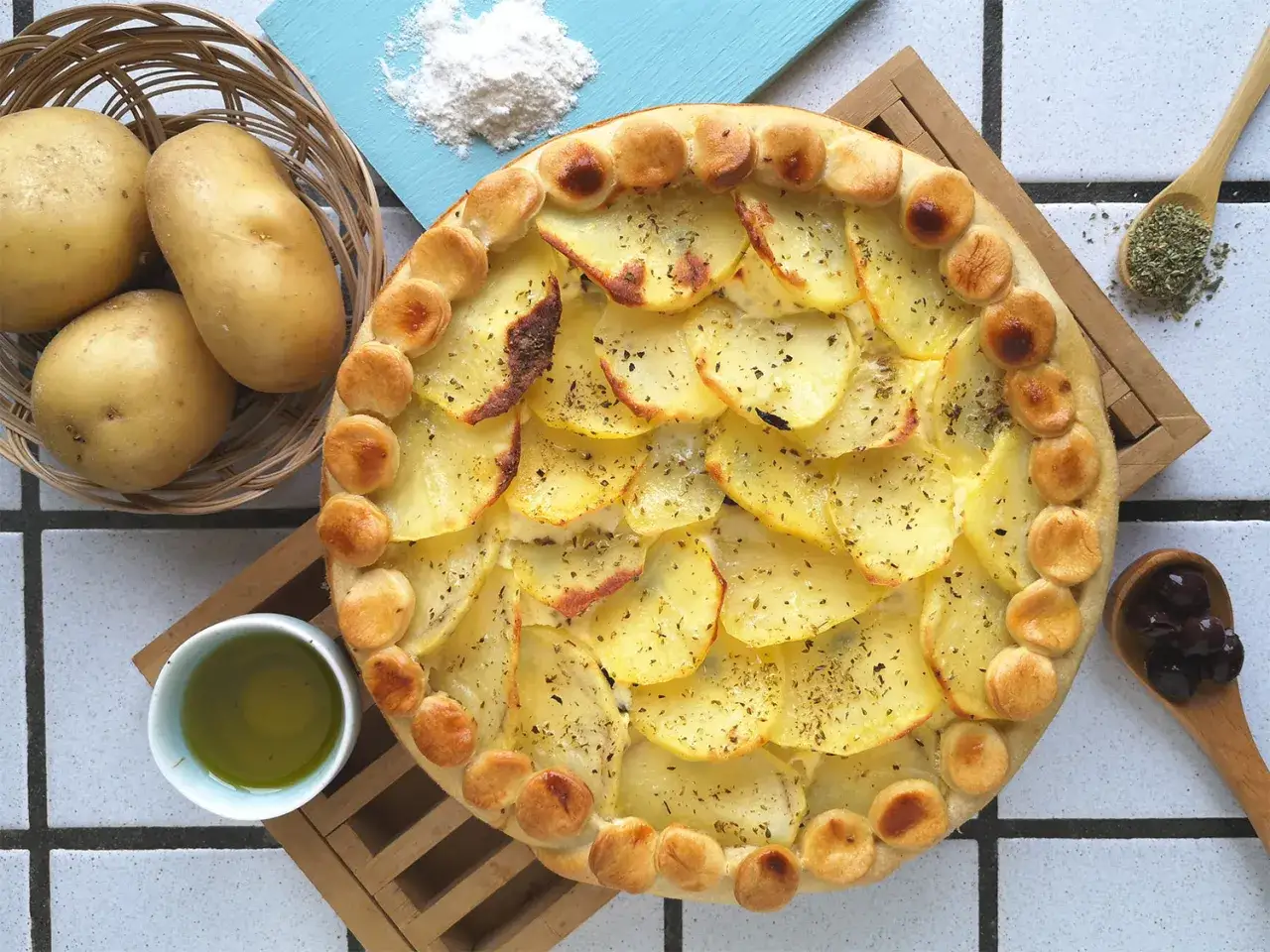 Torta salata con patate, dorata e profumata, con patate affettate e erbe aromatiche. Accanto, patate fresche, olio e olive.