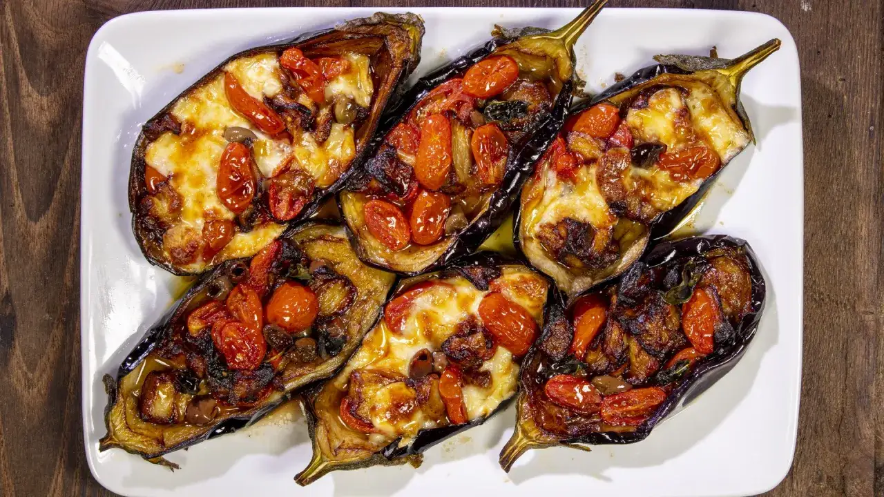 Melanzane a scarpone al forno, ripiene di pomodorini, formaggio filante e olive nere, su un piatto bianco.