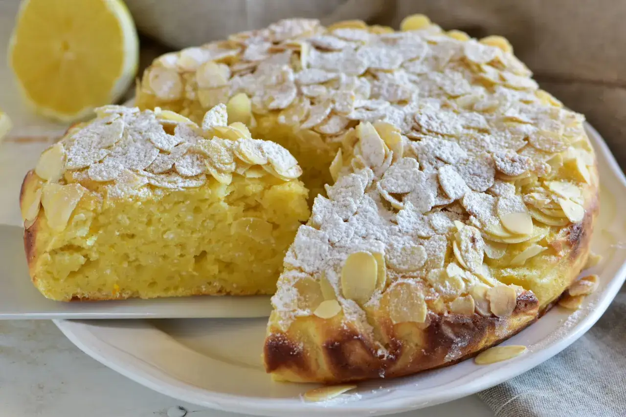 Deliziosa torta con farina di mandorle, cosparsa di zucchero a velo e mandorle a lamelle. Una fetta è già stata servita, mostrando la sua soffice consistenza.