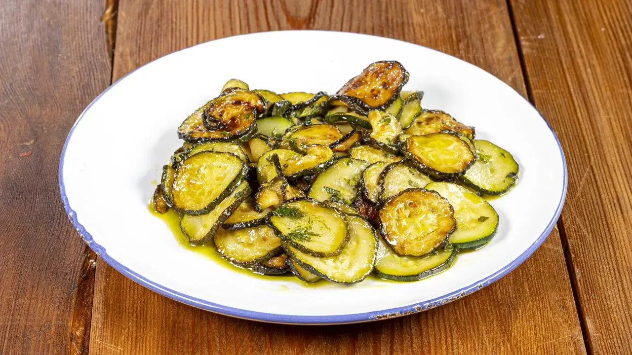 Zucchine alla poverella, fette dorate e succulente condite con olio e erbe aromatiche, servite su un piatto bianco con bordo blu.