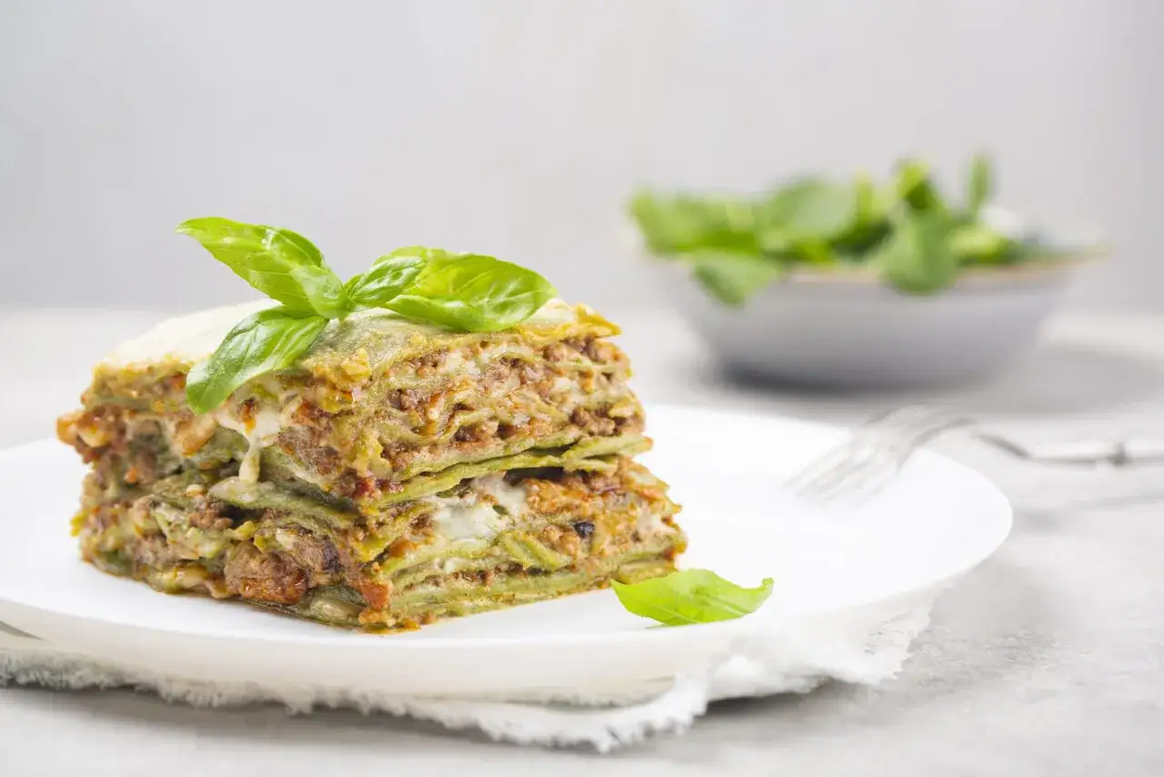 Porzione di lasagne alla bolognese, con strati di pasta verde, ragù e besciamella, guarnita con basilico fresco.