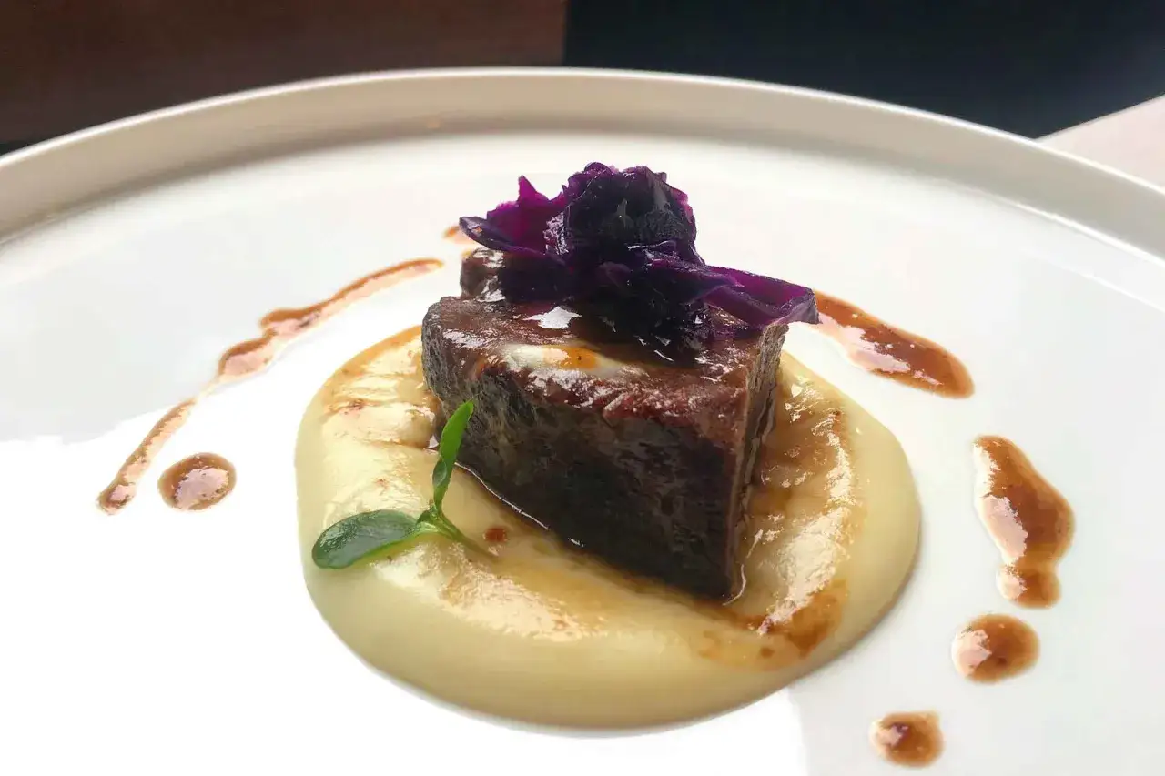 Un succulento cubetto di guancia di manzo tenerissima, adagiato su un letto di purè cremoso, guarnito con un fiore viola e gocce di salsa.