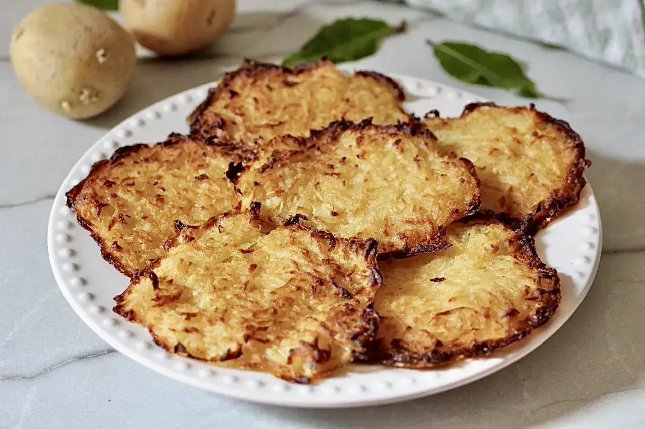 Croccanti rosti di patate dorati su un piatto bianco, con patate fresche sullo sfondo.
