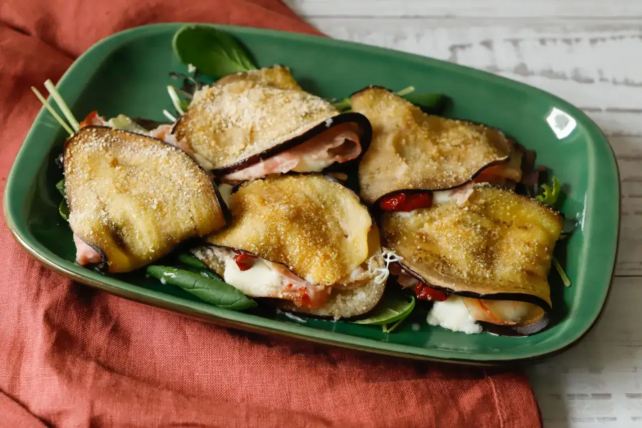 Deliziosi involtini di melanzane impanati, ripieni di formaggio filante e prosciutto, serviti su un letto di insalata fresca.