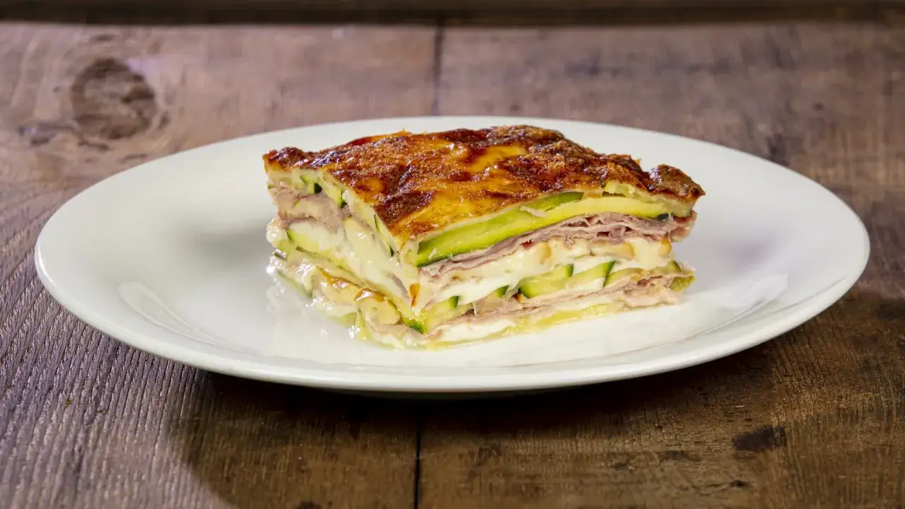 Una fetta di parmigiana di zucchine bianca senza besciamella, con strati di zucchine, prosciutto e formaggio filante, dorata al forno.
