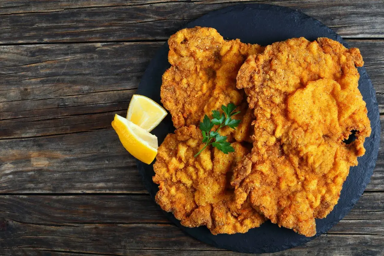 Cotoletta alla milanese dorata e croccante, guarnita con limone e prezzemolo, servita su una pietra scura.