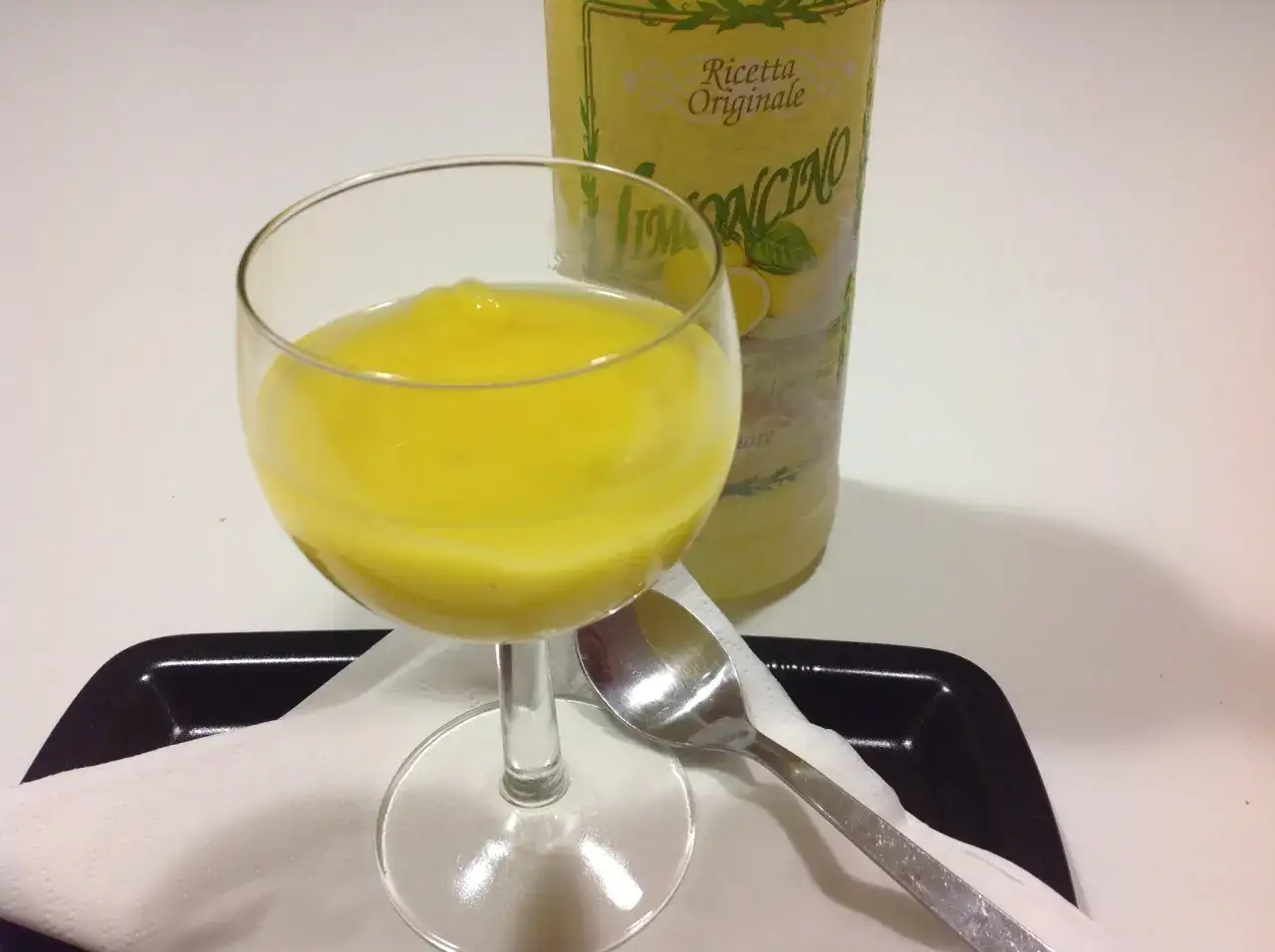 Un bicchiere pieno di crema di limoncello, accanto a una bottiglia. Un cucchiaino è pronto per gustare questo dolce.