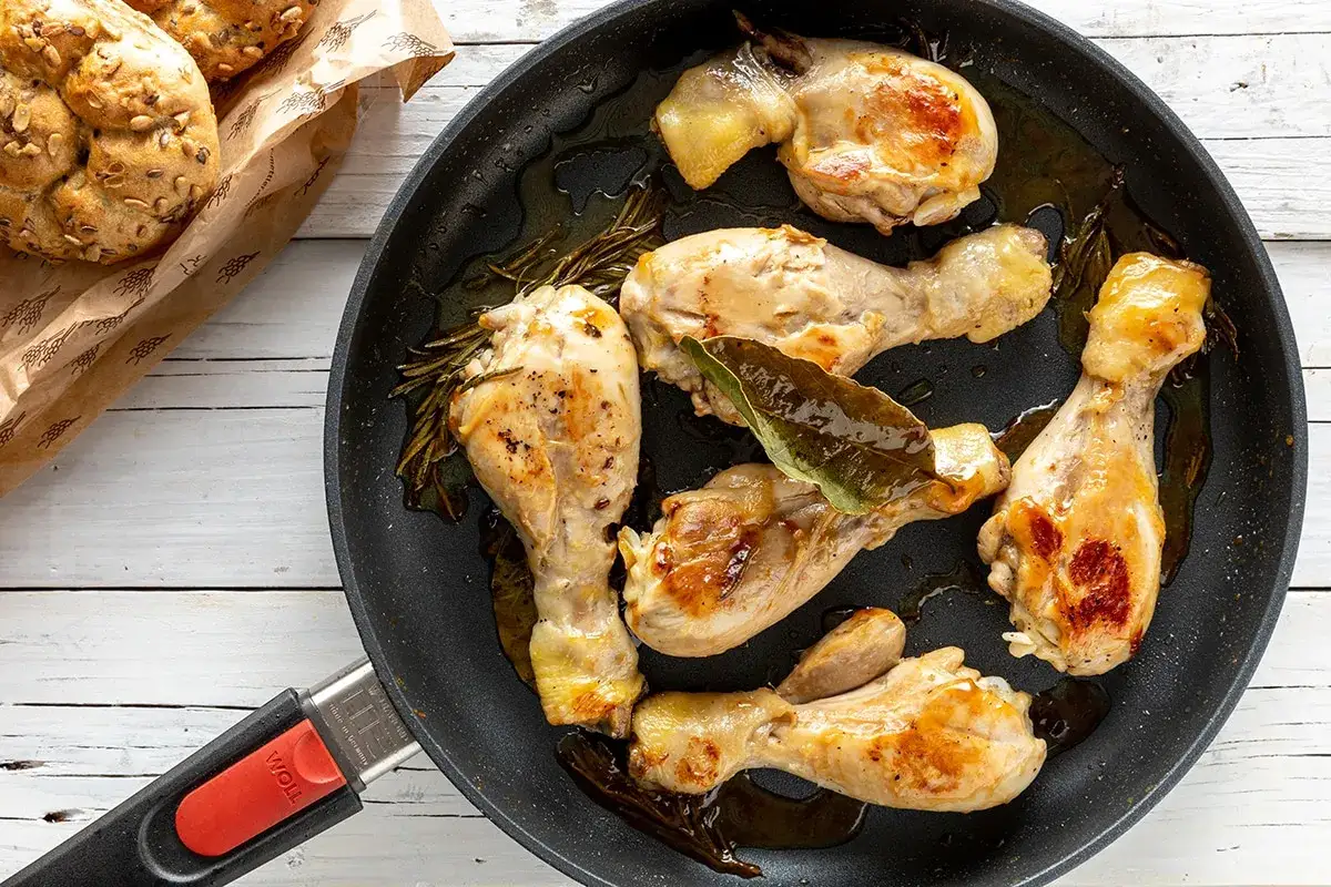 Fusi di pollo in padella, dorati e succulenti, con rosmarino e alloro. Accanto, panini integrali.