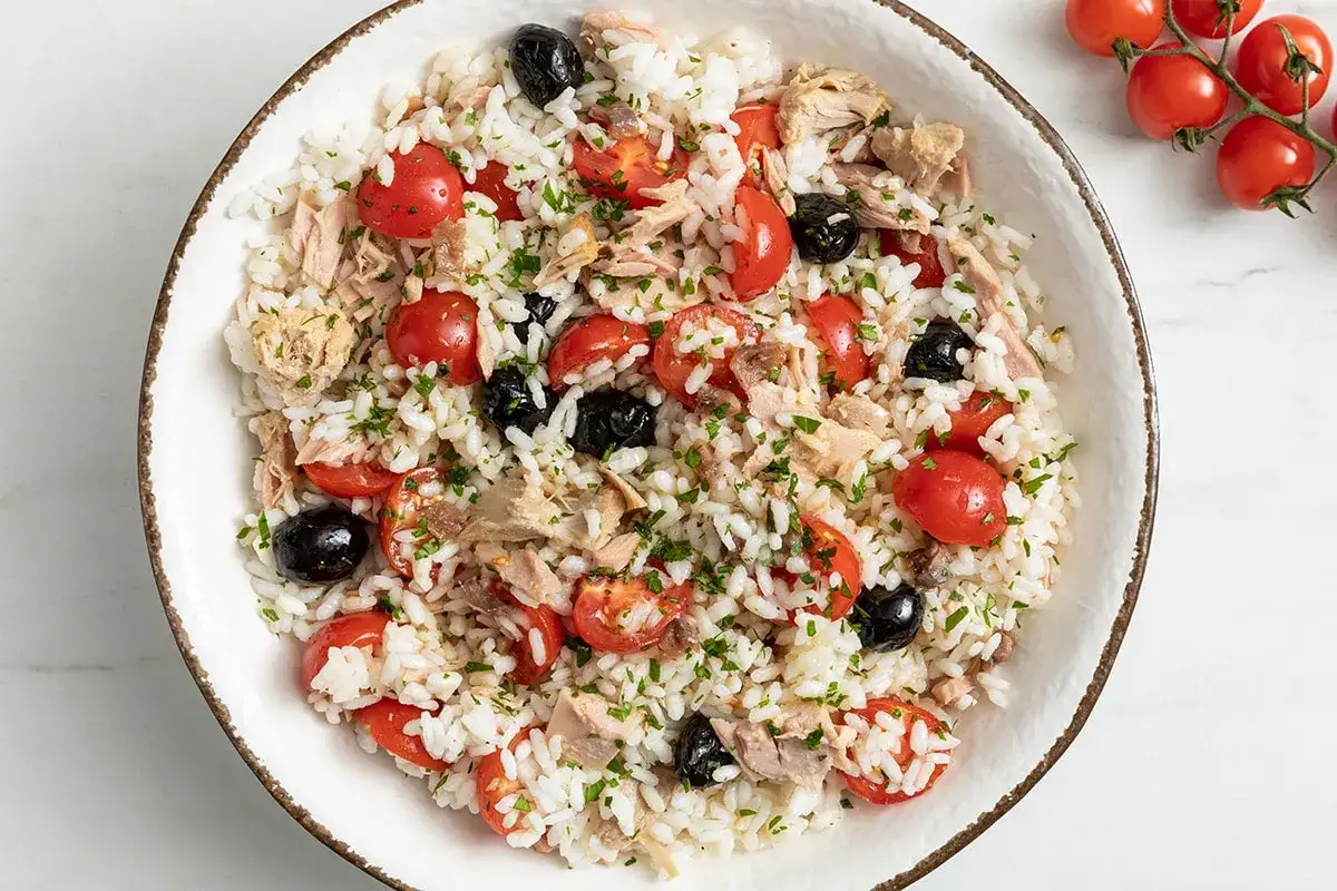 Un'insalata di riso colorata con pomodorini, olive nere e tonno, guarnita con prezzemolo tritato. Perfetta per un pasto leggero e gustoso.