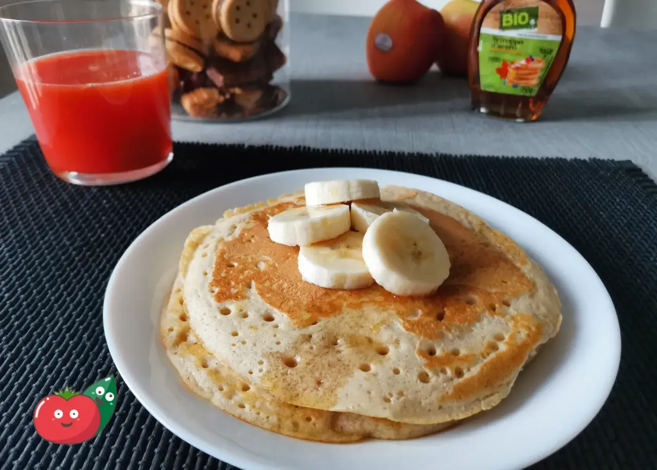 Pancake alla banana dorati, guarniti con fette di banana fresca e sciroppo d'acero. Accanto, un bicchiere di succo rosso e mele.