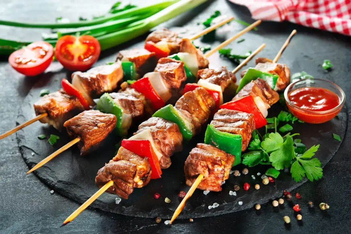 Spiedini di carne succulenti alternati a peperoni rossi e verdi, cipolla bianca, serviti su una pietra scura con pomodorini e salsa.