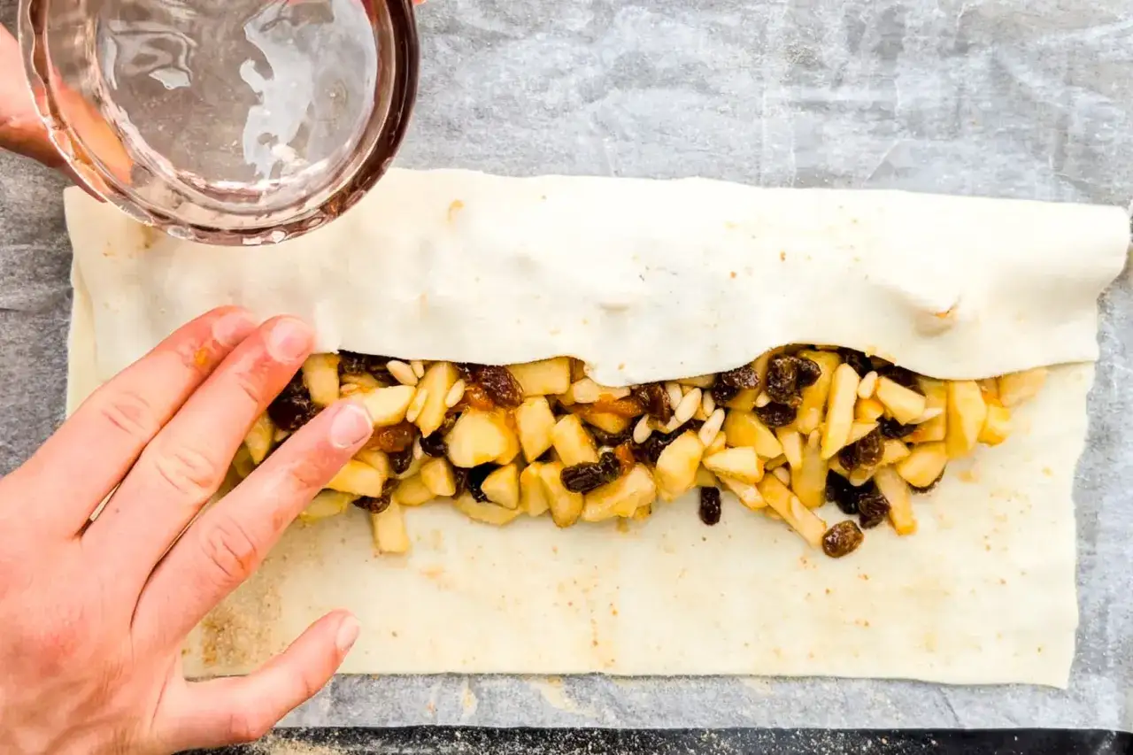 Mani che preparano uno strudel di mele con pasta sfoglia, ripieno di mele a cubetti, uvetta e pinoli.
