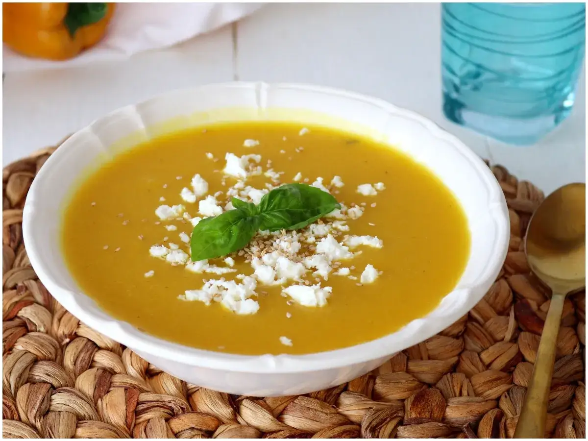 Crema di peperoni gialli, guarnita con feta sbriciolata, basilico fresco e semi di sesamo. Servita in una ciotola bianca su un sottobicchiere intrecciato.