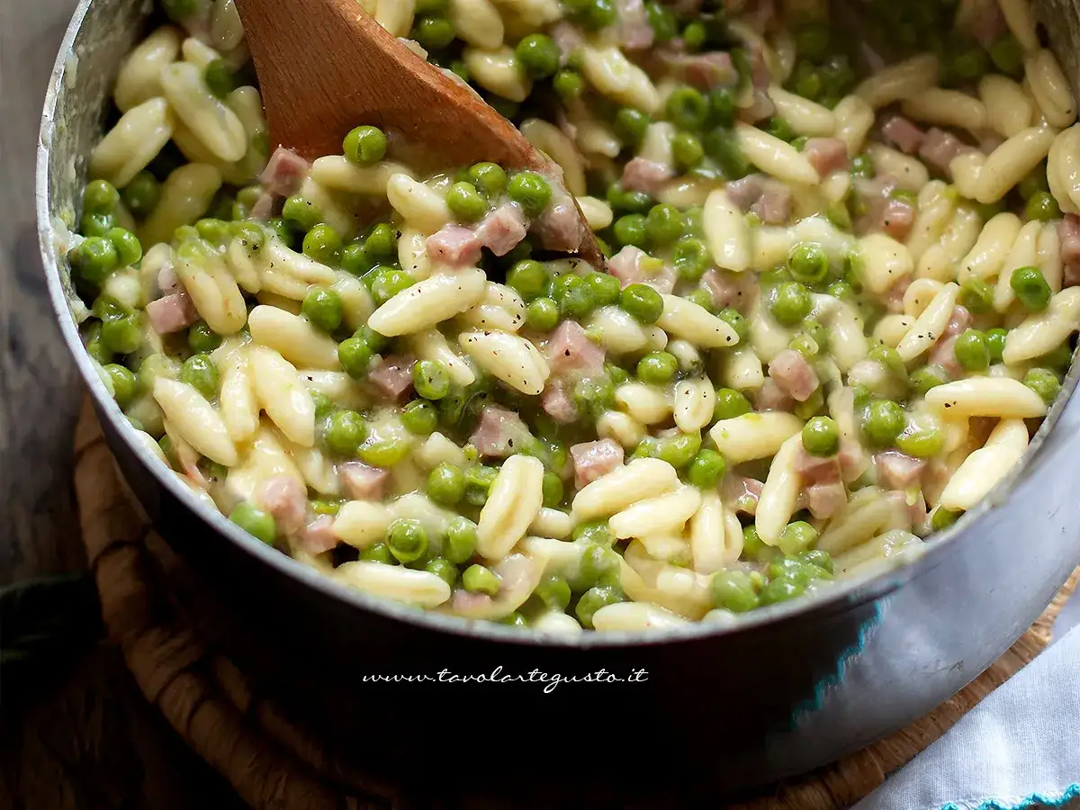 Un primo piatto cremoso: pasta con piselli e cubetti di prosciutto cotto, mescolata con un cucchiaio di legno.